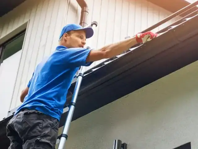 Person on a telescoping ladder reaching into a roof gutter to clear it