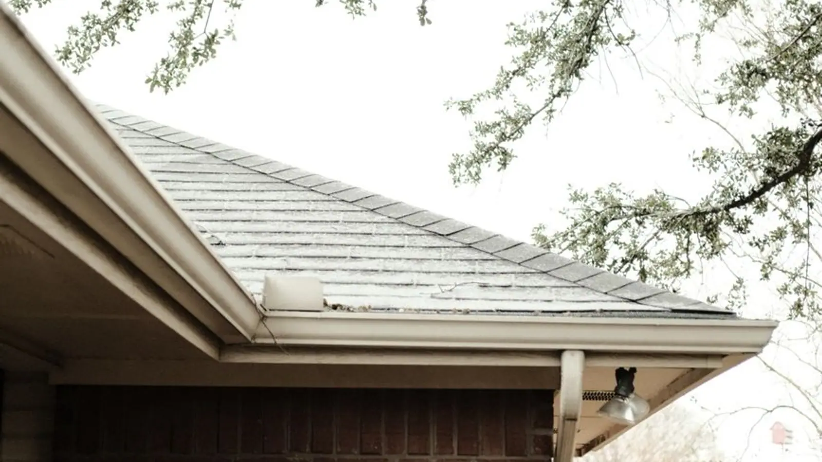 White gutter and downspout along the shingle roof edge of a brick house